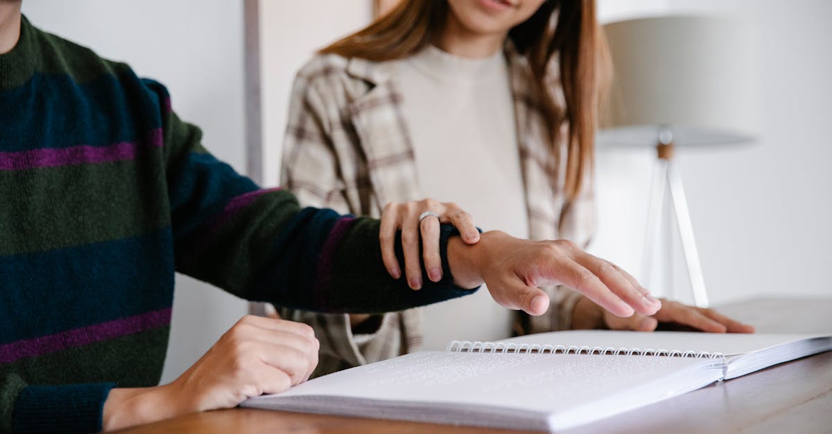 Person guiding another's hand over a Braille book, showing support and assistance indoors.