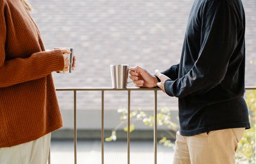 Free Two individuals in casual wear holding mugs while standing on a balcony handrail. Stock Photo