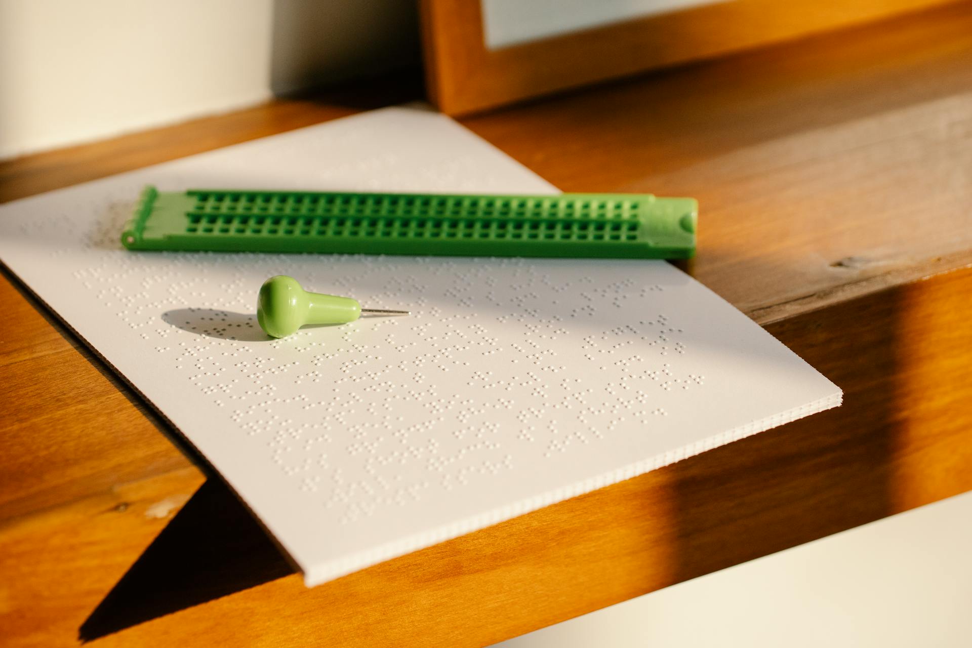 Close-up of braille paper and stylus on a sunlit wooden table, highlighting texture and tools for the visually impaired.