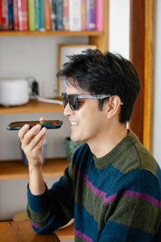 A smiling man wearing sunglasses uses voice command on a smartphone indoors.