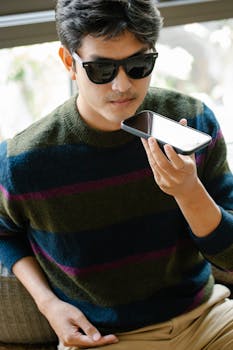 Casual adult male wearing sunglasses using voice command on smartphone indoors.