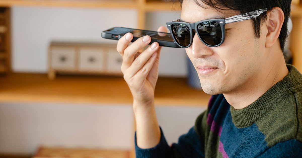 Um homem alegre usando óculos de sol em um ambiente interno, utilizando comandos de voz em seu smartphone.