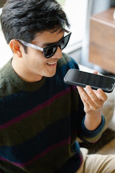 Smiling young man wearing sunglasses, sending a voice message with his smartphone indoors.
