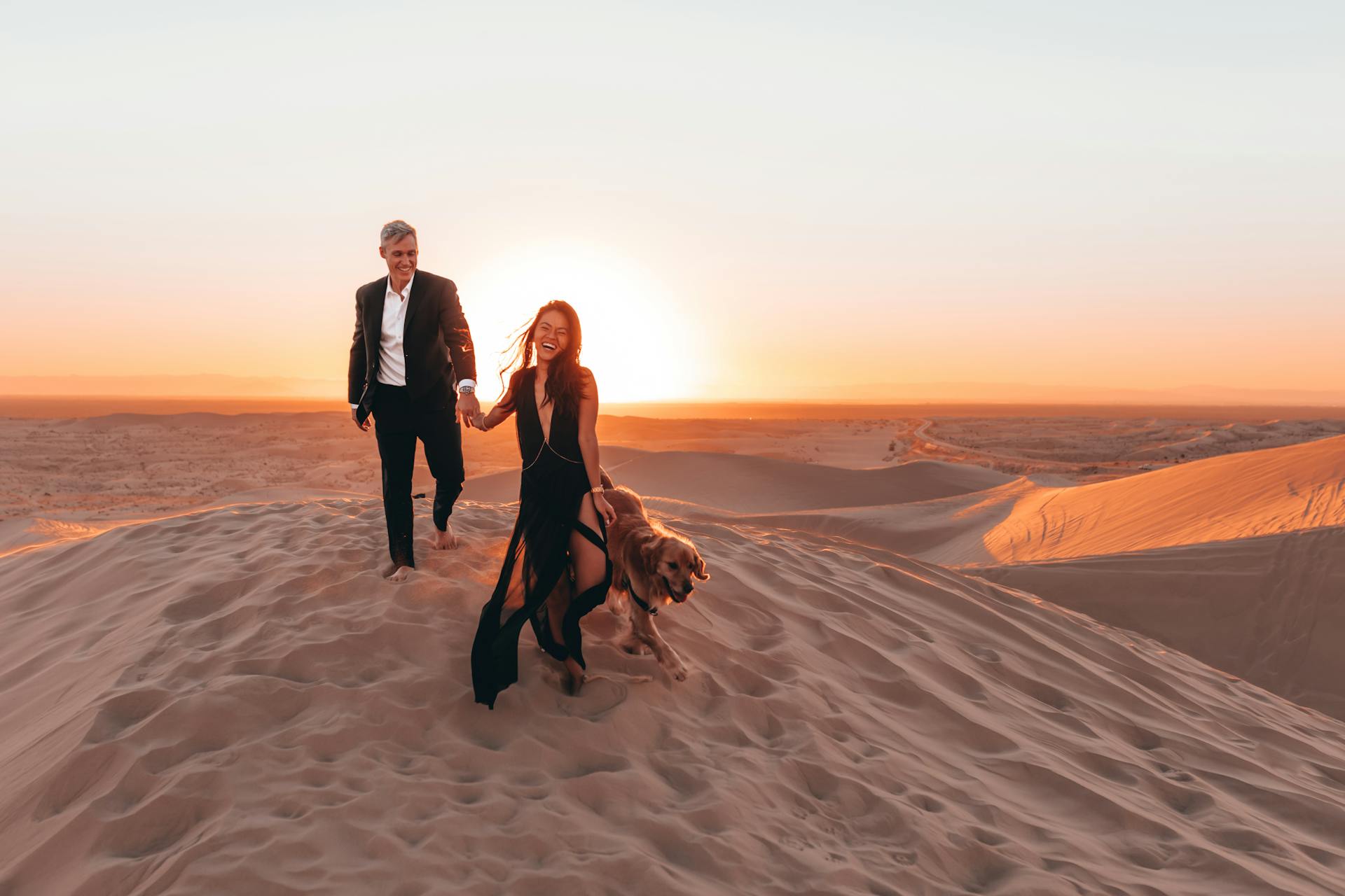 Plein de corps de couple riant positif se tenant par la main et courir avec le chien Golden Retriever sur le sol sableux du désert au soleil