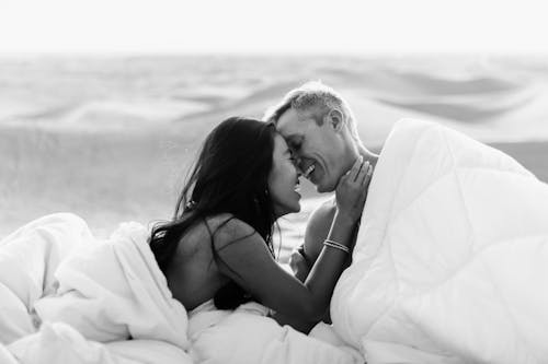 Black and white of positive couple smiling and embracing under white soft blankets on blurred background of desert