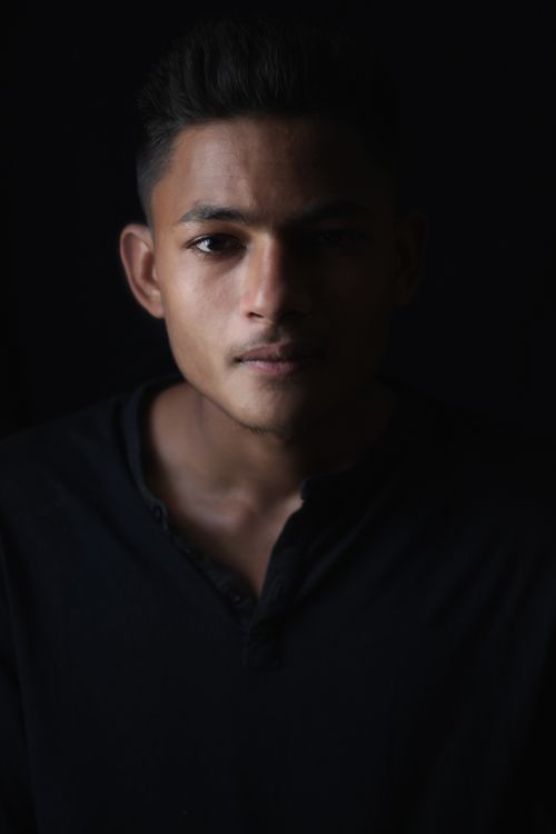 Free Serious Indian man looking at camera with thoughtful gaze while standing in room with dim light and shadow covering half face on black background Stock Photo