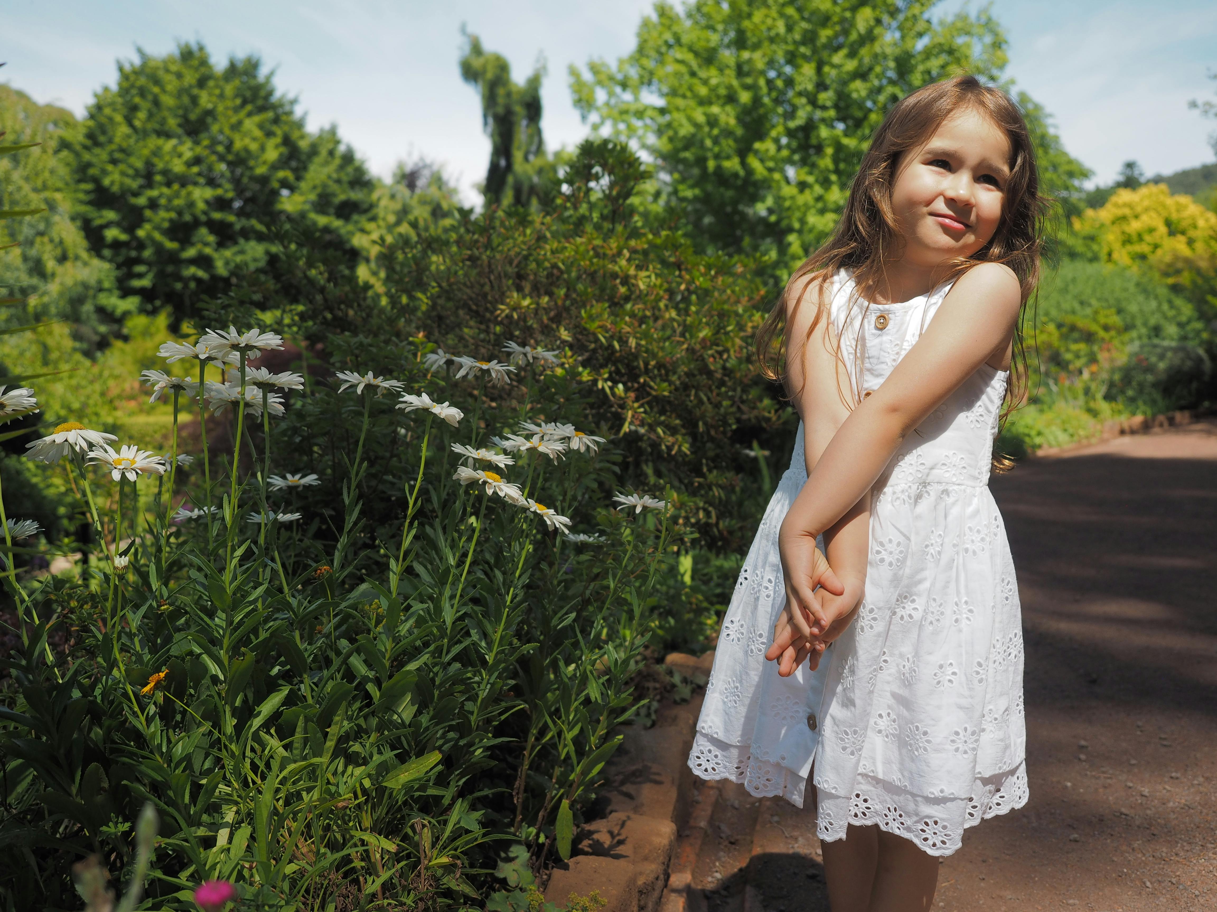 A young girl in a white dress shyly poses in a vibrant garden filled with white flowers on a sunny day.