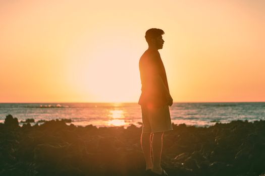 A serene silhouette of a man standing on a rocky beach, backlit by a stunning ocean sunset.