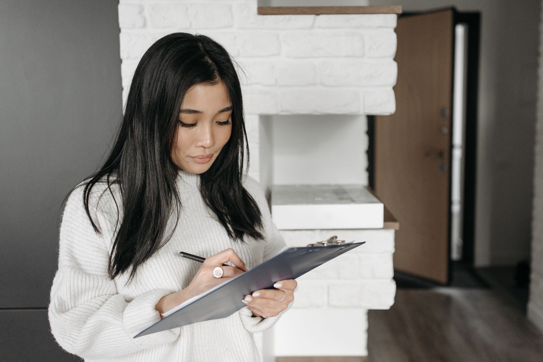 Asian woman focused on writing on a clipboard indoors. Casual, at-home setting.