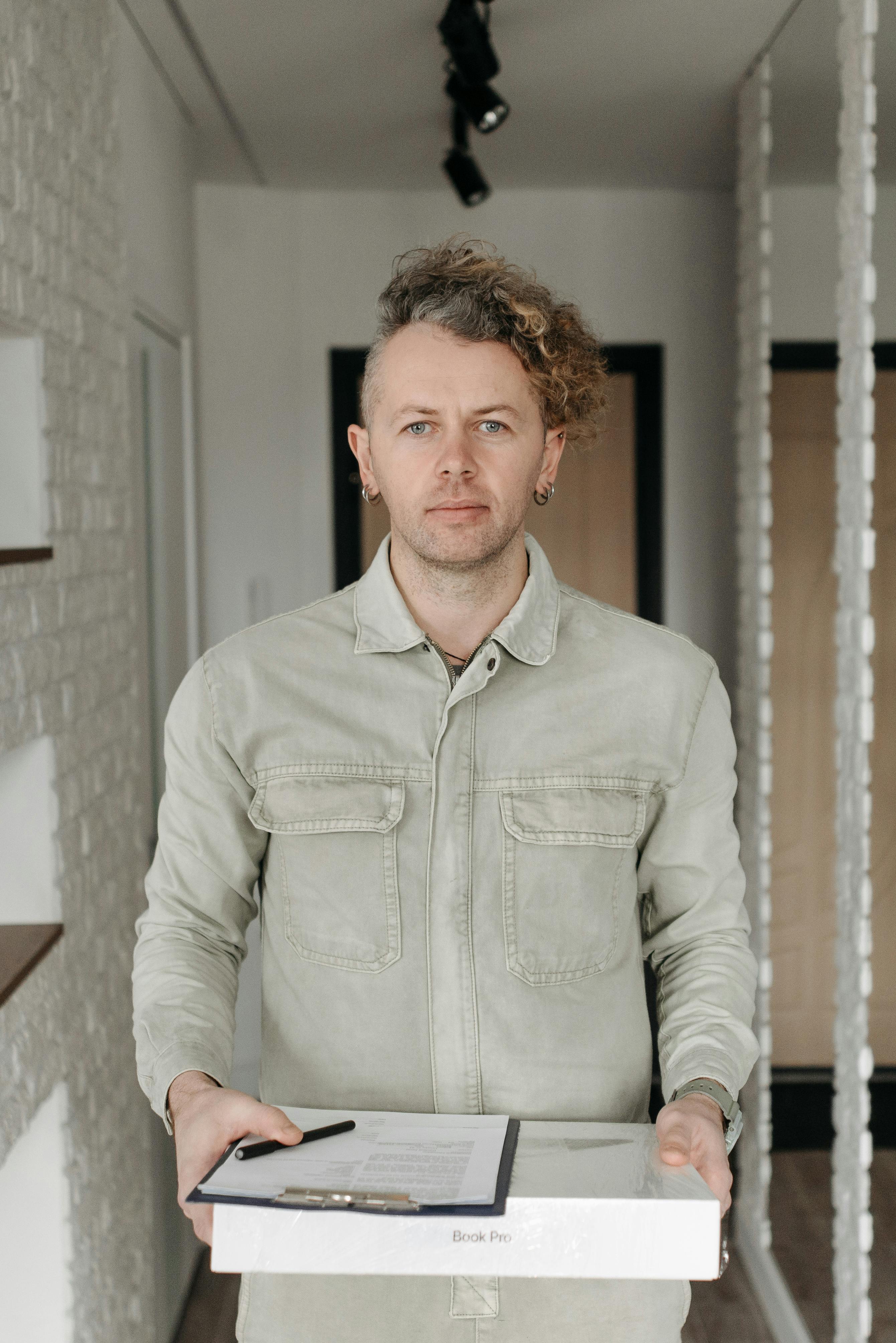 Young adult man holding a package and clipboard inside a contemporary home setting, symbolizing online delivery services.