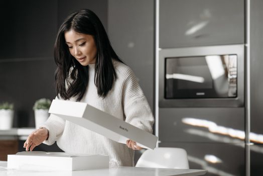 An asian woman excitedly unpacks a delivery box in her modern kitchen.