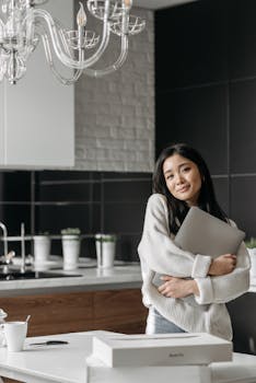 Smiling Asian woman holds laptop in stylish kitchen, illustrating comfort and technology.
