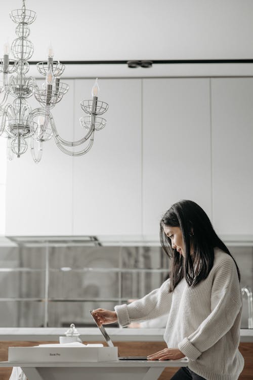 Free Woman in White Sweater Looking at the Laptop While Standing in the Kitchen Under Chandelier  Stock Photo
