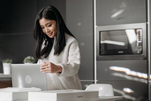 Free An Asian woman smiling as she works on her laptop in a sleek, modern kitchen setting. Stock Photo