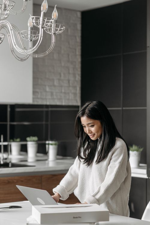 Free Smiling Asian woman using a laptop in a stylish, bright kitchen setting. Stock Photo