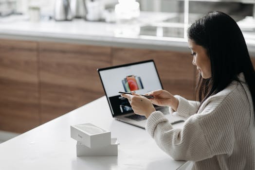 Young Asian woman setting up a new smartphone while working on a laptop in a modern indoor setting.