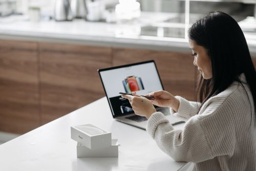 Free Young Asian woman setting up a new smartphone while working on a laptop in a modern indoor setting. Stock Photo