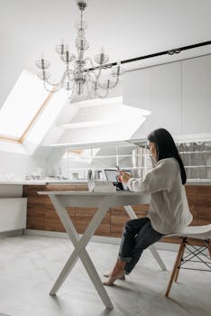 A woman sits in a bright, modern kitchen using a laptop and mobile phone, embracing a digital nomad lifestyle.