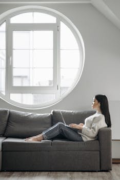 Young woman in a sweater using a laptop on a comfortable sofa near a round window.