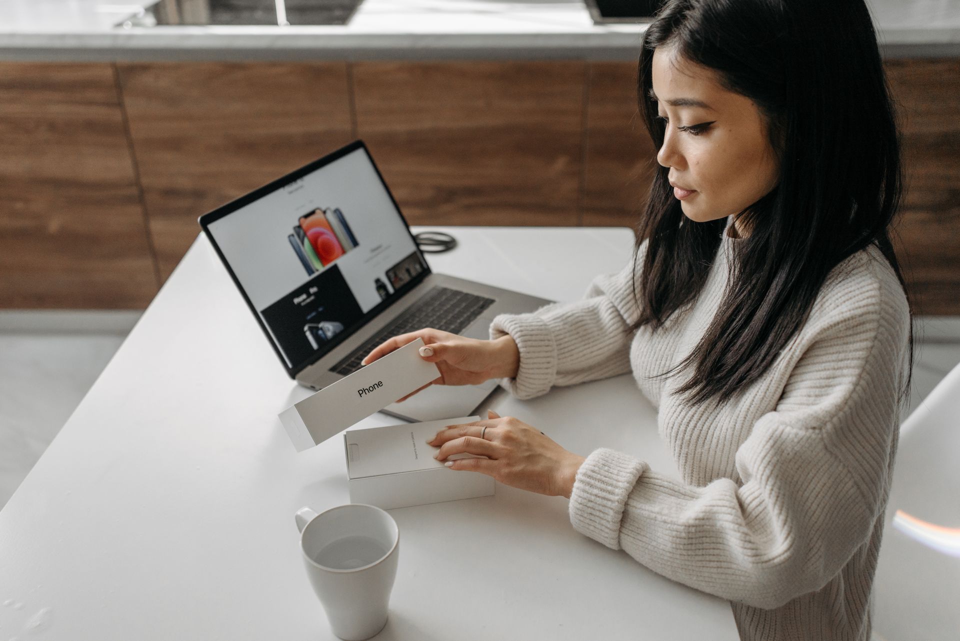 Adult woman unboxing a smartphone with a laptop nearby, showcasing technology lifestyle.
