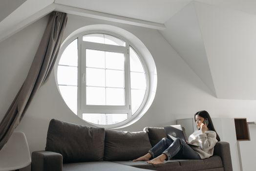 Asian woman sitting on a sofa, working from home on a laptop while on a phone call.