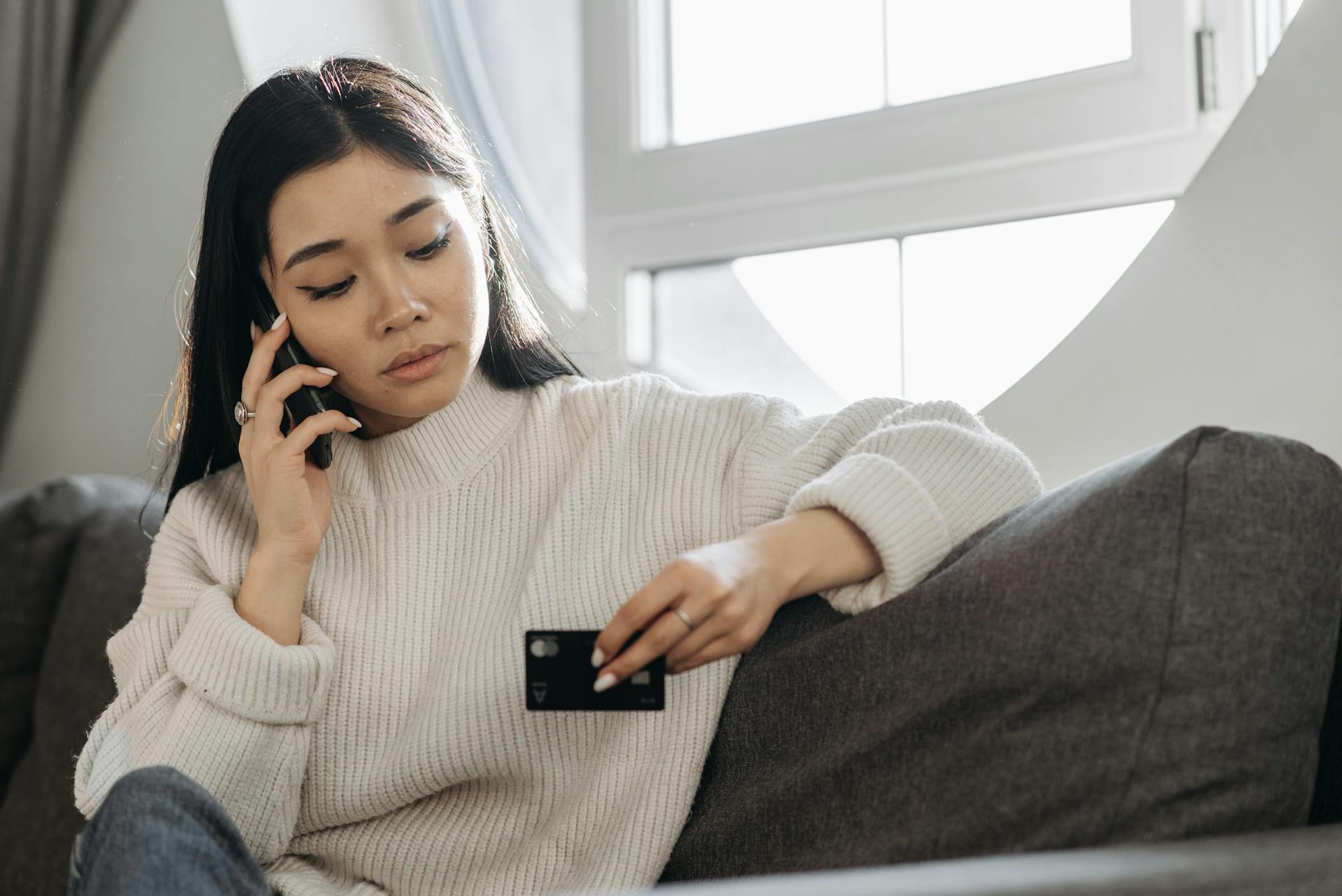 A woman sits on a sofa, multitasking with a phone and credit card, focusing on online shopping.