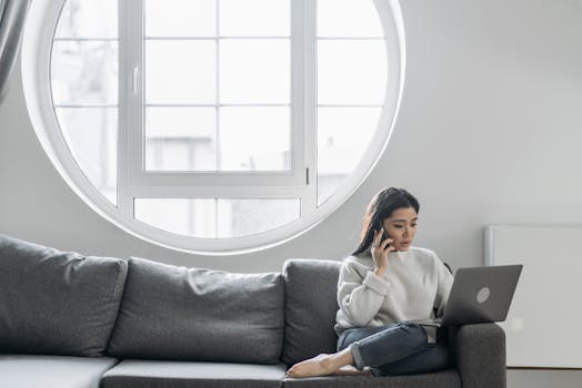 Asian woman multitasking at home with a laptop and phone, sitting on a sofa.