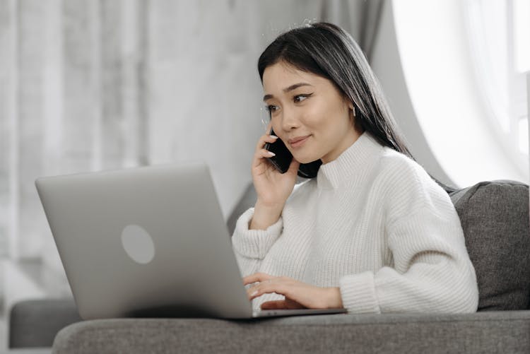 Woman In White Sweater Talking On Phone While Using Laptop