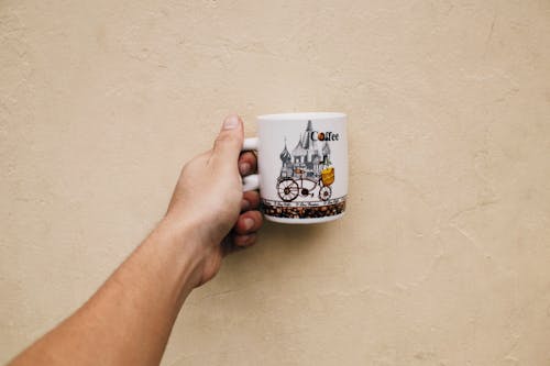 Free Crop anonymous male demonstrating ceramic mug with picture for coffee near white wall Stock Photo