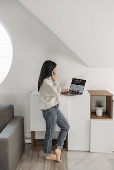 A woman stands indoors wearing a sweater, using a laptop for online shopping.