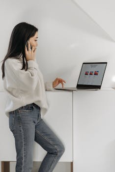 Modern asian woman multitasks using a laptop and smartphone indoors in a minimalist setting.