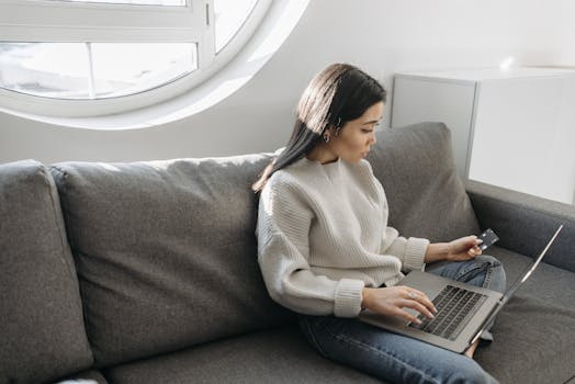Woman sitting on a sofa using a laptop for online shopping indoors, holding a credit card.