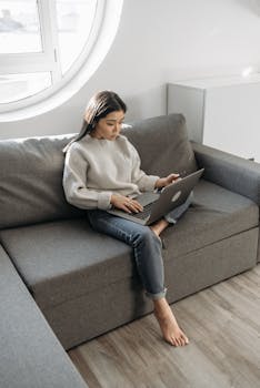 Asian woman sitting on a couch, working on a laptop in a well-lit room.