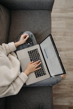 Top view of a woman shopping online using a laptop while sitting on a grey sofa.