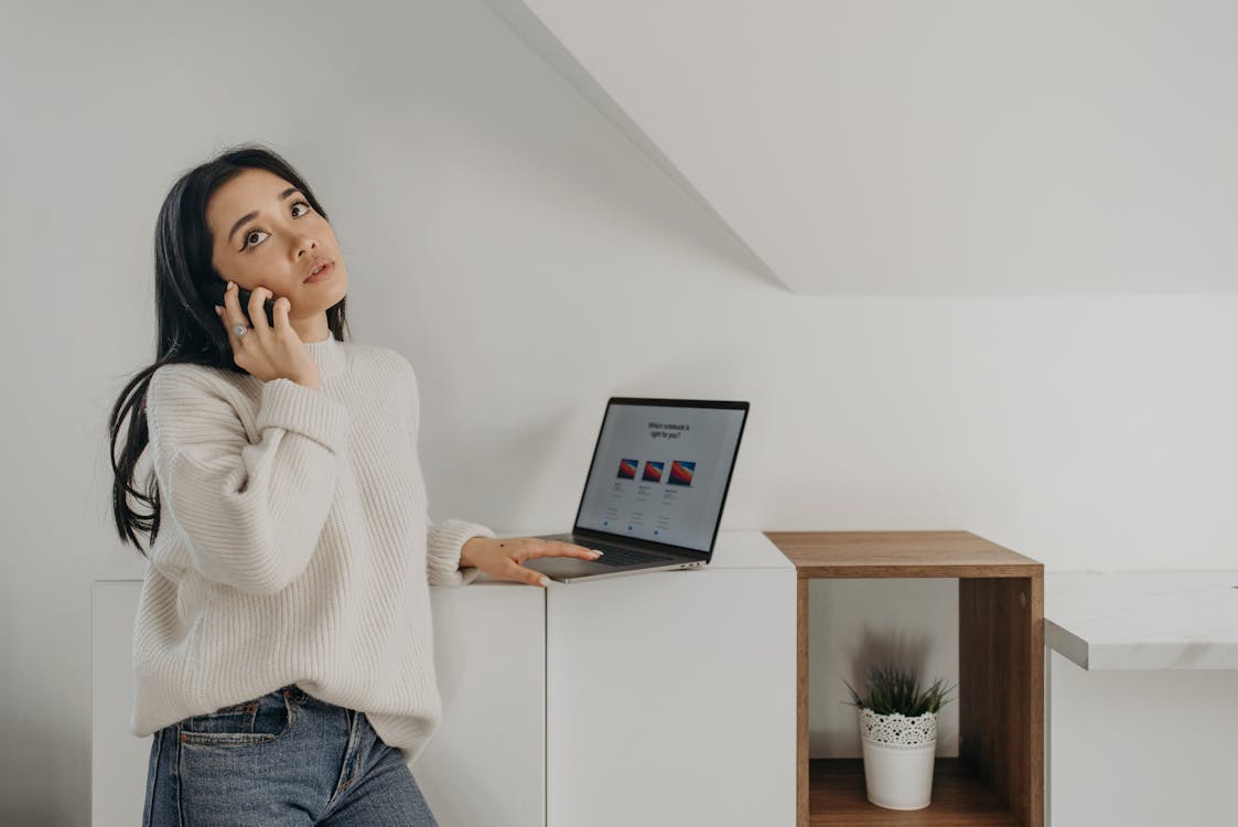 Free Woman in White Sweater Talking on Phone while Using Laptop Stock Photo