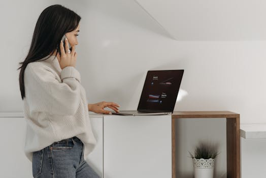 Asian woman multitasking with a mobile phone and laptop in a bright indoor setting.