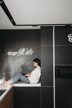 Adult woman using laptop while sitting on a countertop in a contemporary kitchen setting.