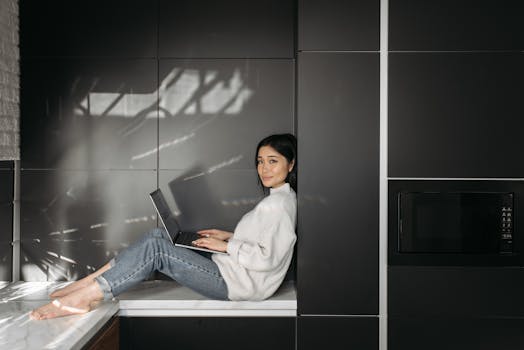 Young woman sitting on countertop using laptop for remote work in stylish kitchen.