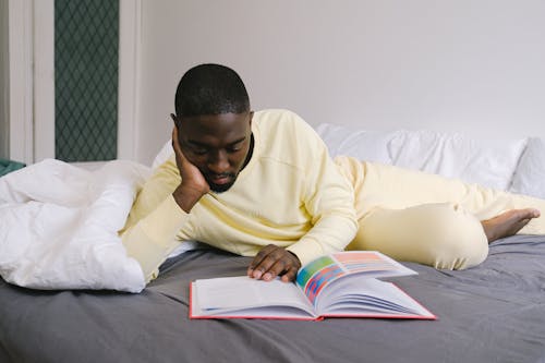 Free Man in Yellow Sweater Reading a Book on Bed Stock Photo