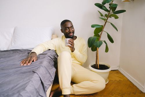 Free Relaxed man enjoying a morning coffee sitting on the bedroom floor next to a plant. Stock Photo
