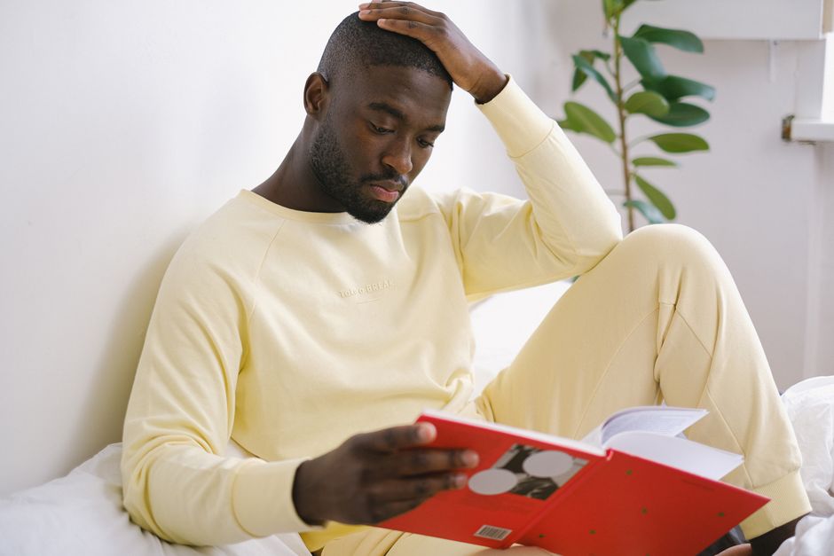 Pensive African American man in pajamas with workbook touching head while preparing for exam on bed in house Pensive African American man in pajamas with workbook touching head while preparing for exam on bed in house