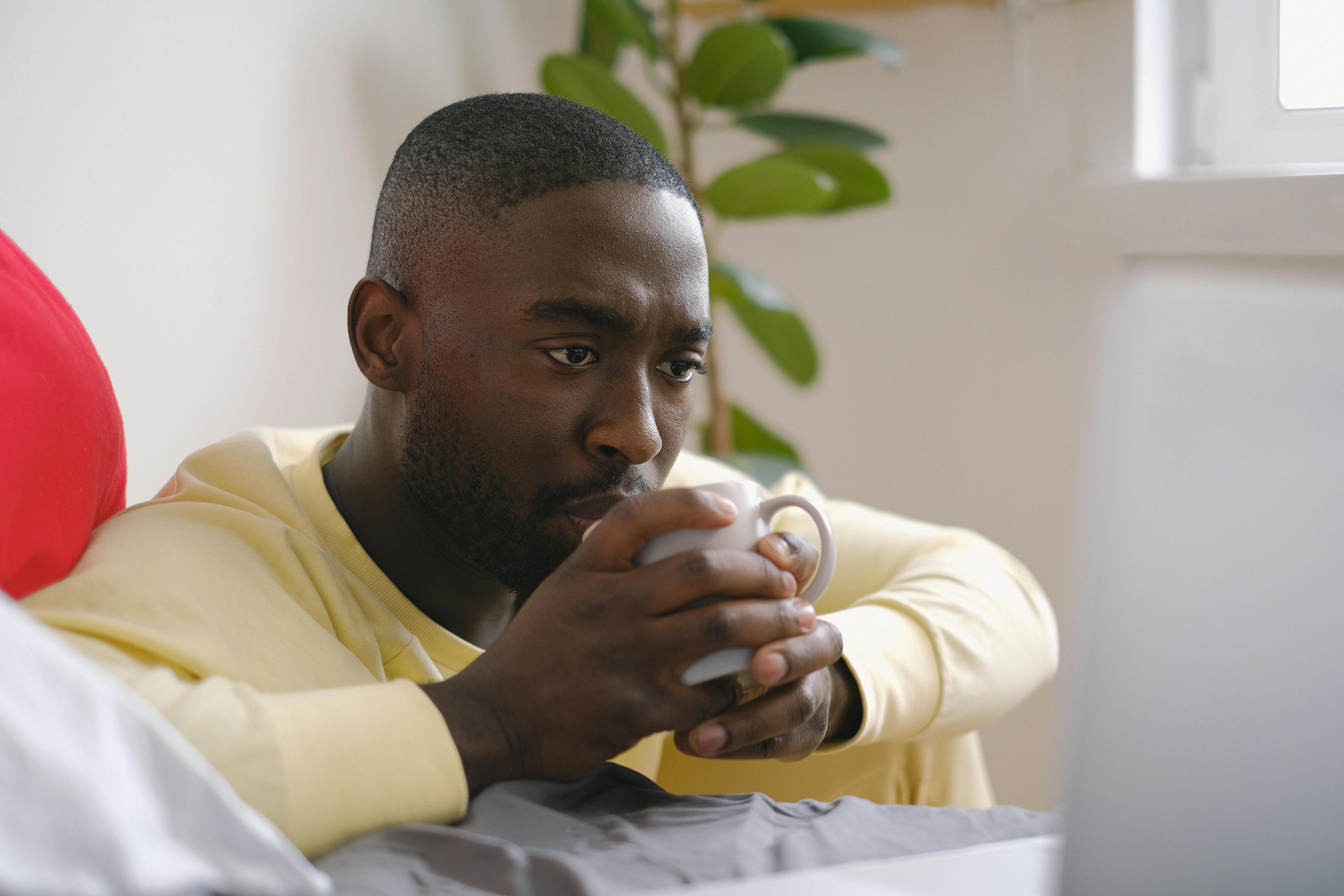 attentive black man drinking coffee against laptop on bed