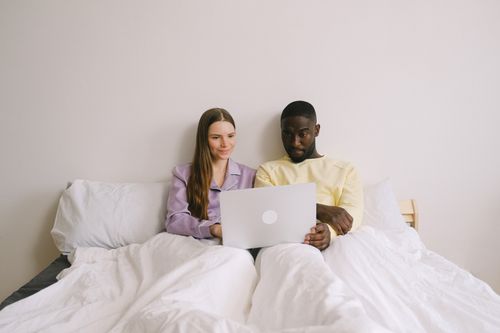 Free Interracial couple sitting in bed using a laptop, showcasing love and technology at home. Stock Photo