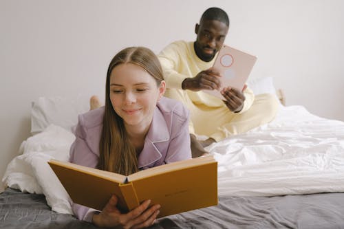 Free Cheerful woman with book lying on soft bed near positive African American boyfriend browsing modern tablet in light bedroom at home Stock Photo
