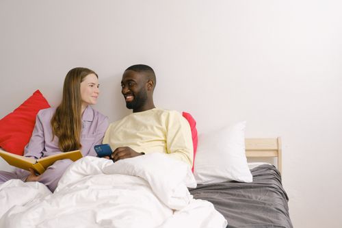 Free Cheerful woman with notebook and positive African American man with cellphone looking at each other while resting on bed in bedroom Stock Photo