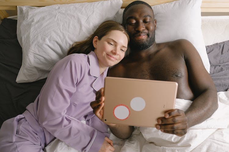 Cheerful Diverse Couple Browsing Tablet On Bed