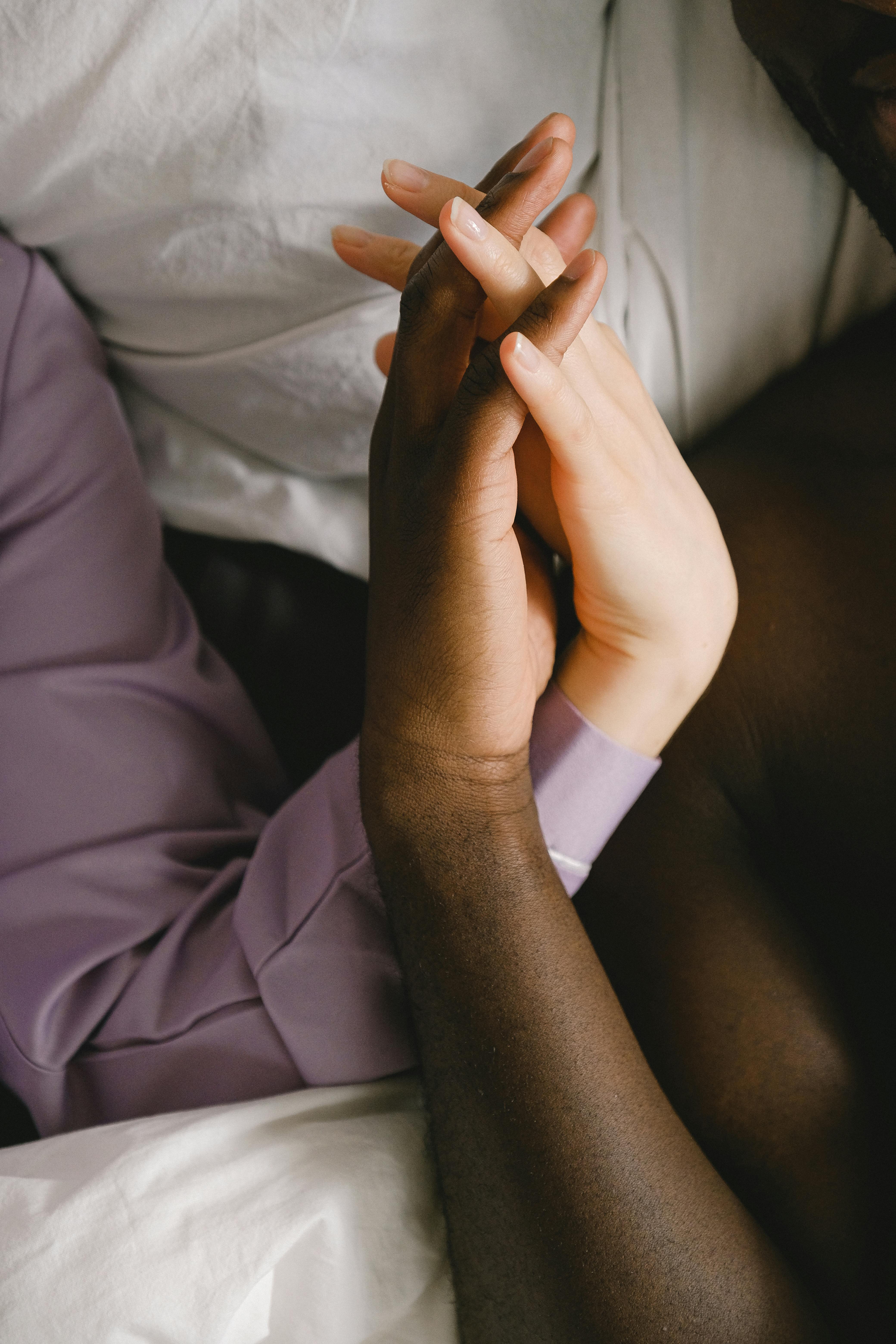 Top view of crop anonymous diverse loving couple holding hands while resting on bed together with white bedclothes in bedroom