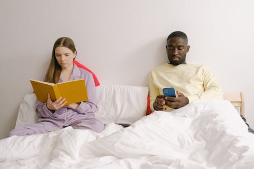 Focused diverse couple with book and smartphone on bed