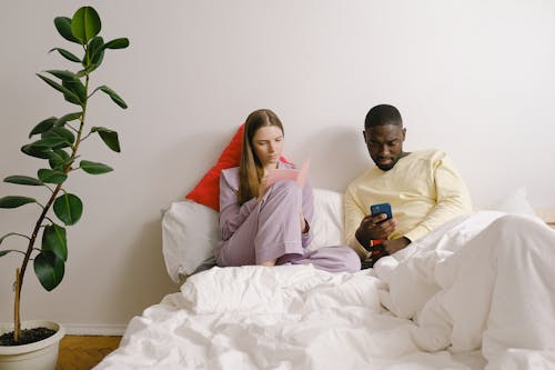 Free Focused woman taking notes in notebook near African American man surfing cellphone while sitting on bed in bedroom with green potted plant Stock Photo