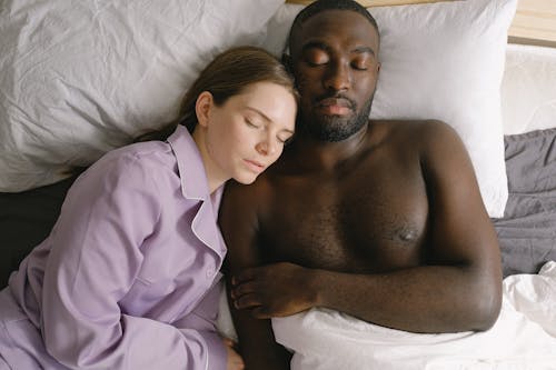 Top view of woman in nightwear and African American man sleeping together on comfortable bed in light bedroom at home